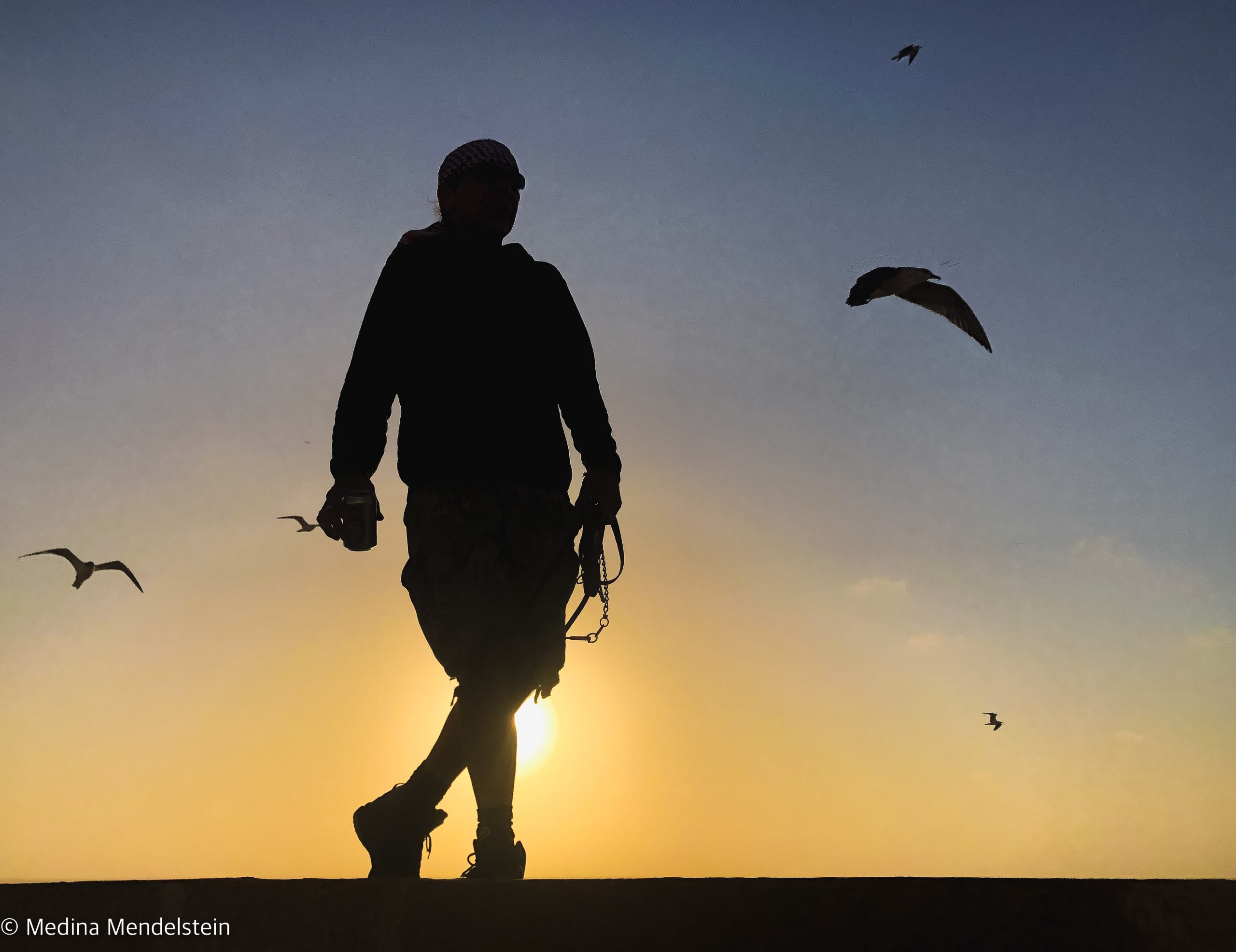 Marokko, Essaouira: Ein Mann tanzt bei Sonnenuntergang auf einer Mauer. Im Hintergrund fliegen Vögel.