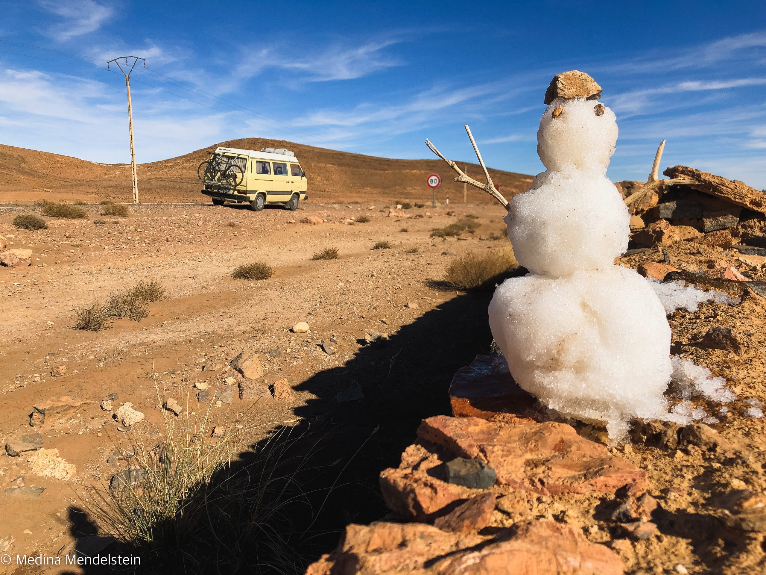 Camping Marokko im Atlasgebirge: Im Vordergrund ein Schneemann auf einer Mauer. Im Hintergrund Wüste und das Atlasgebirge. Auf der Straße steht ein VW T3 Wohnmobil.
