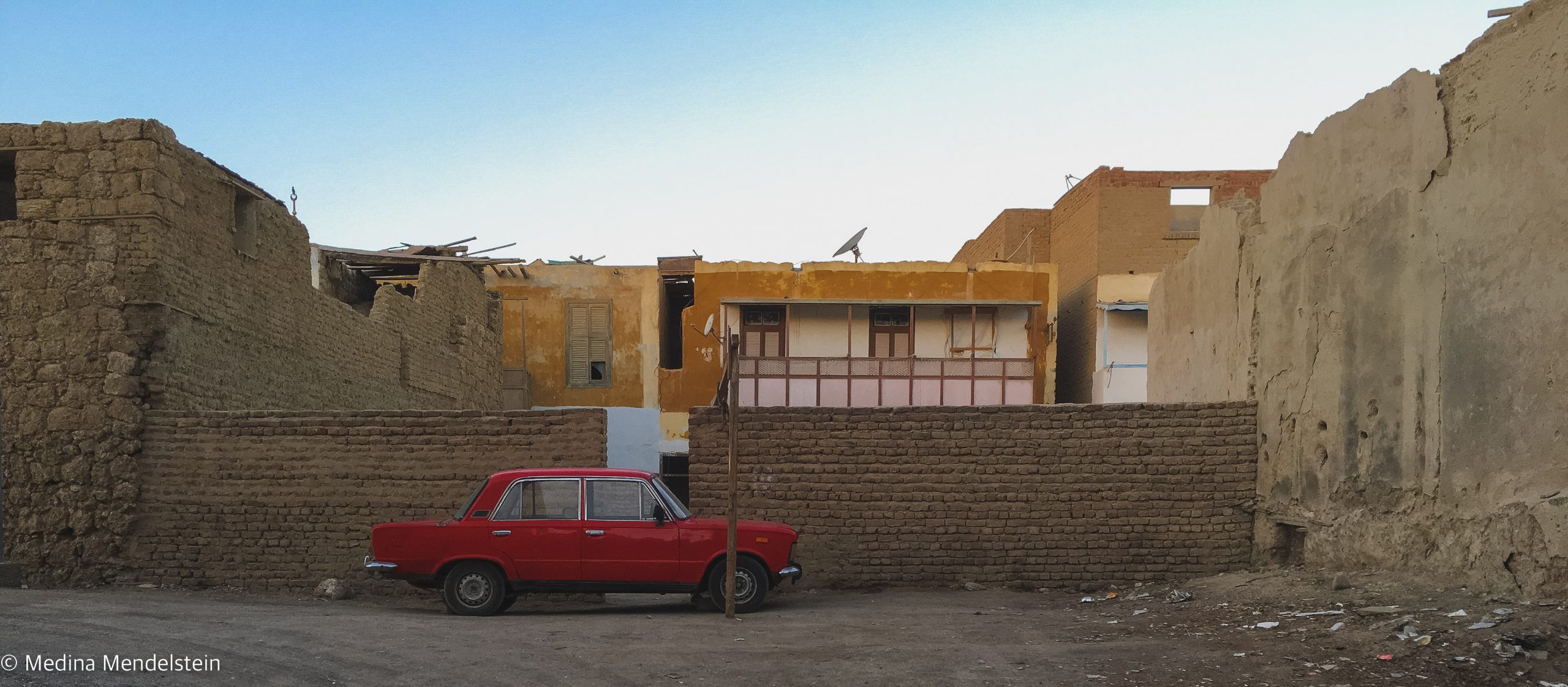 Fotografie aus: al-Quasai in Ägypten, Afrika: Dorf mit Lehmziegelhäusern. Vor den Häusern ist eine Lehmziegelmauer, davor steht ein rotes Auto.