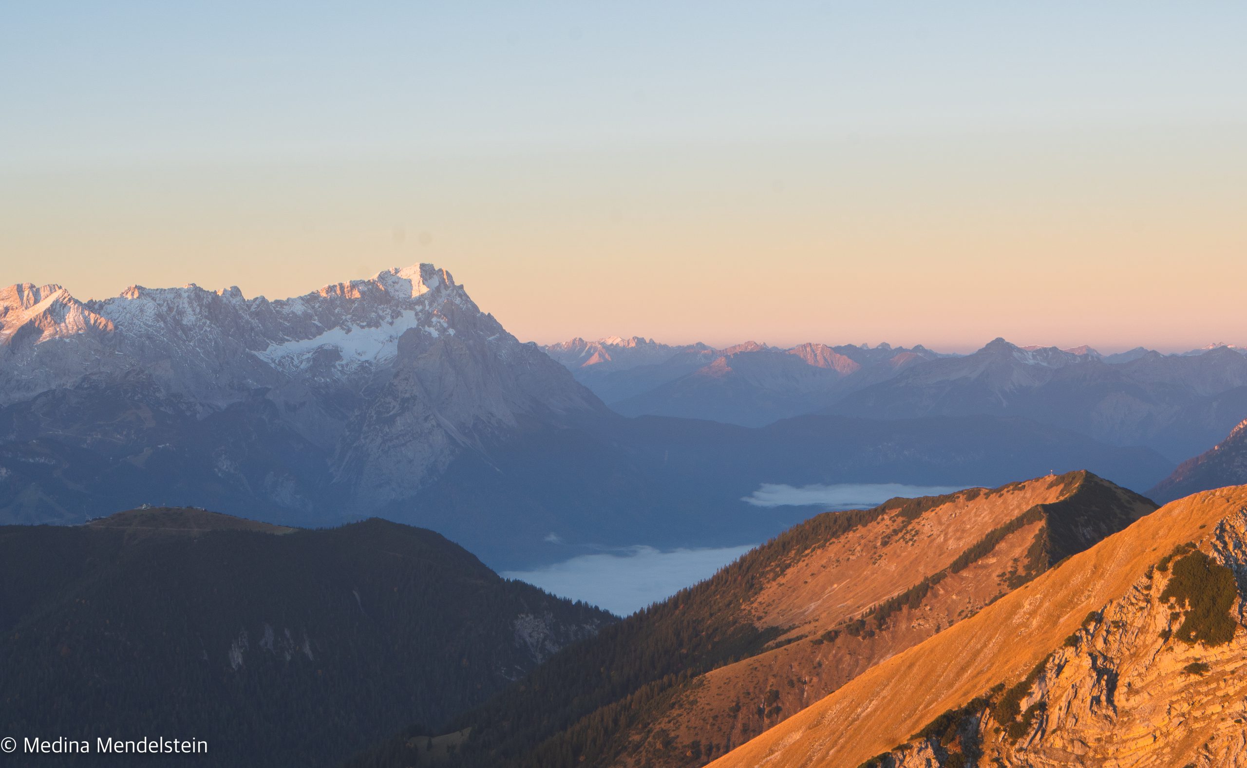 Alpenglühen im Estergebirge. Im Vordergrund ist ein Berghang, der vom Sonnenlicht rot gefärbt ist. Im Hintergrund ist eine Bergkette, die leicht mit Schnee bedeckt ist. Der Himmel ist rot-blau.