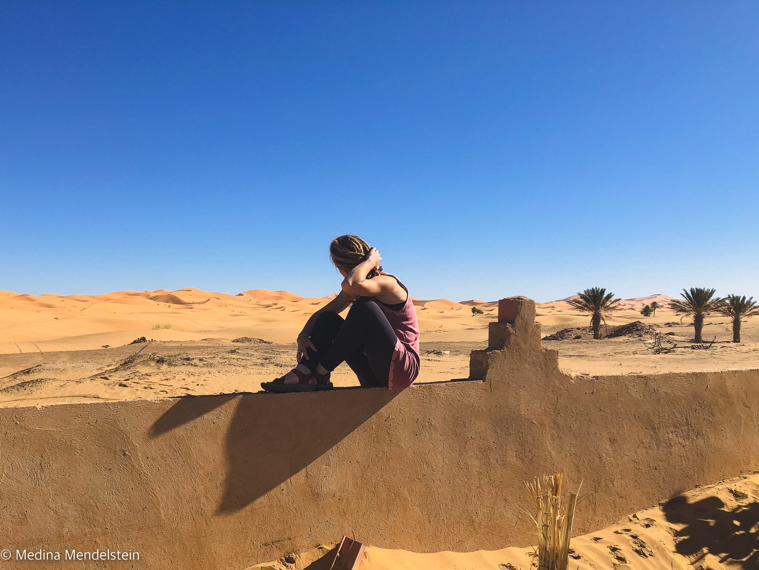 Fotografie aus: Merzouga in Marokko, Afrika. Im Hintergrund ist Marokkos Wüstendüne. Im Vordergrund ist eine Mauer, auf dieser sitzt Medina Mendelstein, Content Creator.