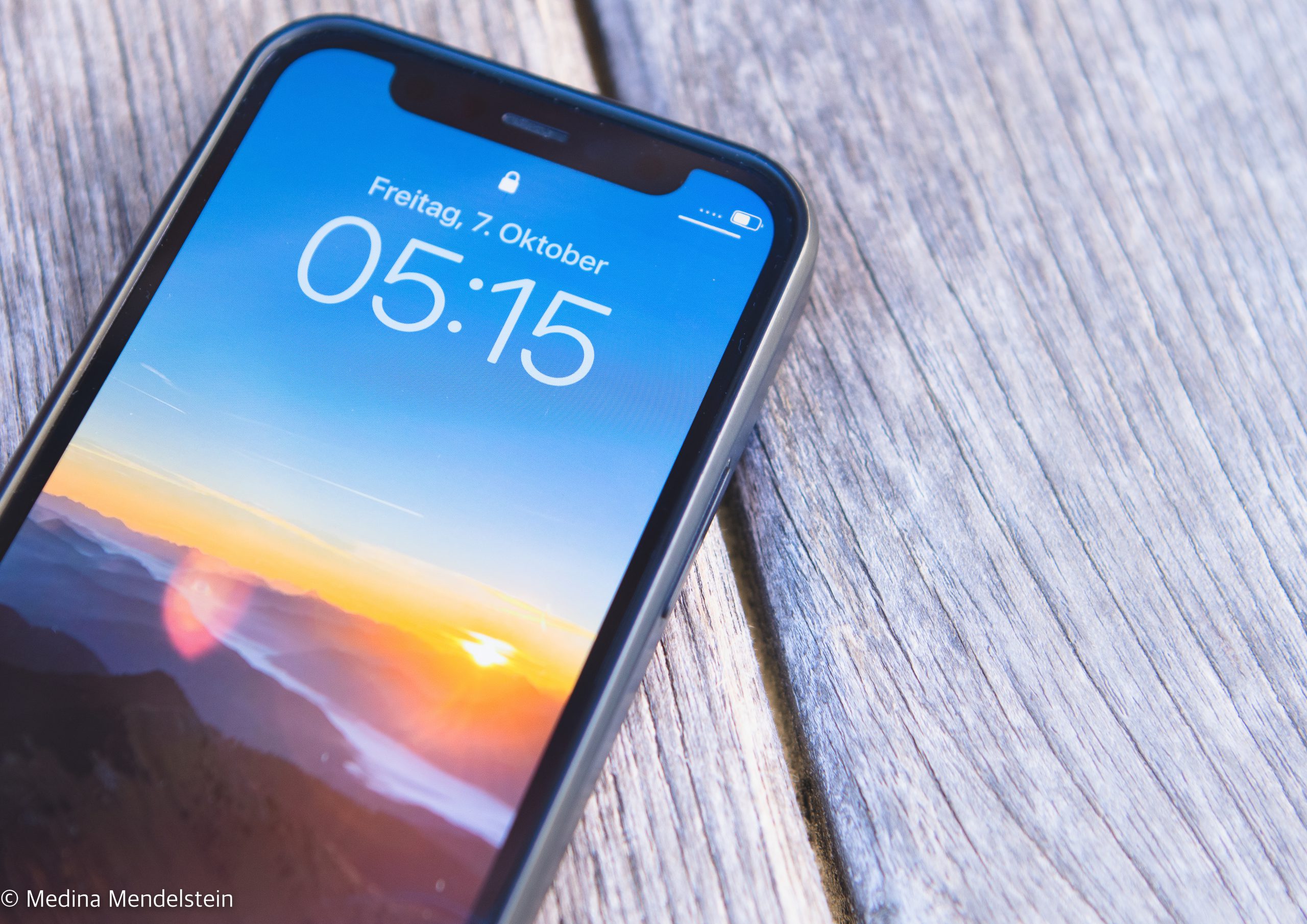 Ein Smartphone, das die Uhrzeit 05:15 Uhr an einem Freitag anzeigt. Das Handy hat kein Netz und Internet Empfang. Das Hintergrundbild des Smartphones zeigt einen Sonnenaufgang im Estergebirge in den Alpen.