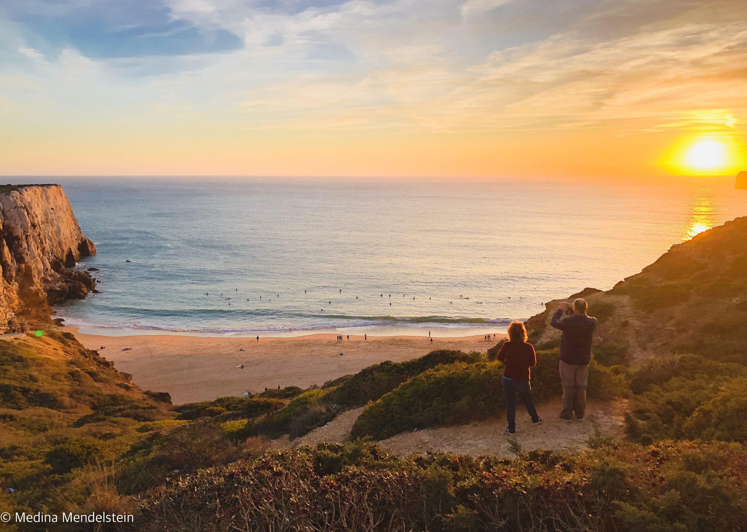 Fotografie aus: Sagres in Portugal, Europa: Strand bei Sonnenuntergang von einer Steilküste aus fotografiert. Im Wasser sind viele Surfer. Auf einem Vorsprung stehen zwei Menschen und schauen zum Wasser.
