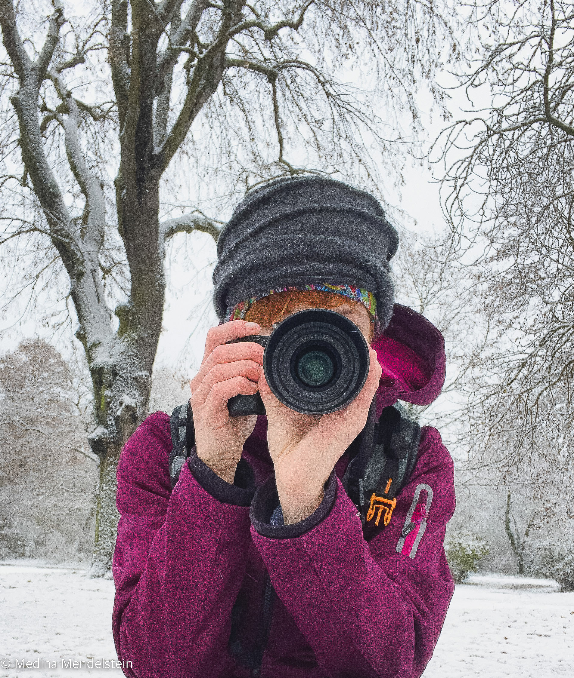 Fotografie – Medina Mendelstein, Content Creator, mit einer Sony Alpha Kamera in der Hand. Die Kamera ist auf den Betrachter des Fotos gerichtet. Im Hintergrund liegt Schnee.